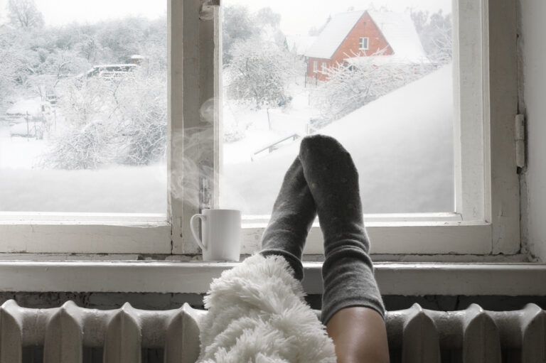 woman legs in warm socks under blanket and mug of hot drink on windowsill, snow outside, prepared for winter storm